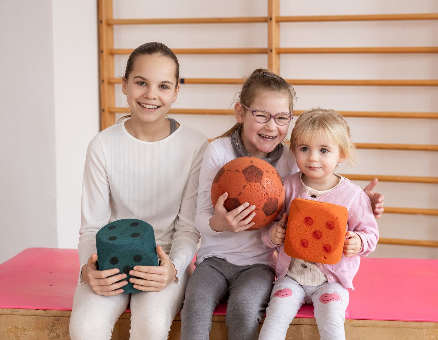 Drei Mädchen mit bunten Softbällen sitzen lachend in einer Turnhalle.