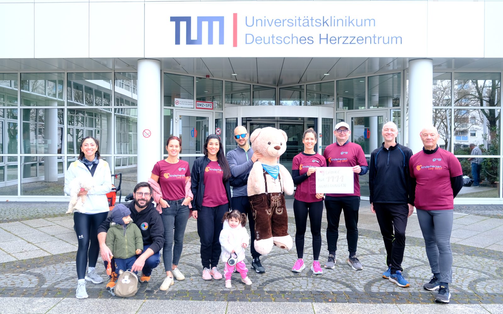 Kinderherzenlauf 2025: Läuferinnen, Läufer und Kinder in den Shirts des Laufs stehen mit einem großen Teddybären und einem Dankesschild vor dem Deutschen Herzzentrum München.